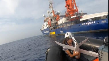 Das Bild zeigt eine Person, die eine Drohne hält, in einem Schlauchboot das die im Hintergrund liegende "Sea-Watch 4" ansteuert.