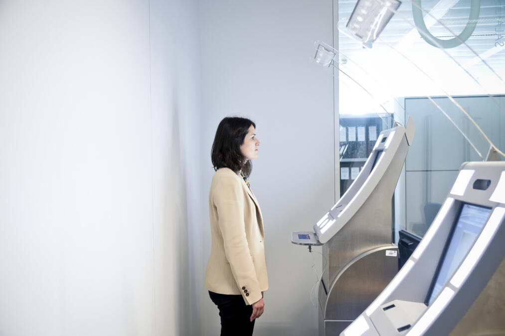 Eine Frau steht aufrecht vor einem Selbstbedienungskiosk mit einem Bildschirm, offenbar wird dabei ihr Gesicht gescannt.