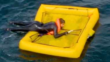 Das Bild zeigt eine gelbe, quadratische Rettungsinsel im Wasser, darauf eine liegende Person.