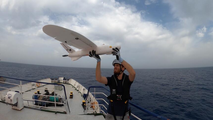 Das Bild zeigt einen Mann, der eine weiße Drohne in der Hand hält und dabei auf einem Schiff im Meer steht.