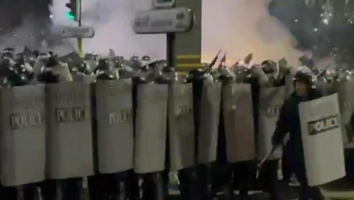 Polizisten mit Schilden und Schlagstöcken bilden eine Reihe, im Hintergrund Rauchschwaden.