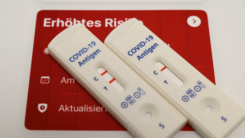 Rote Warnung der Corona-Warn-App mit Schriftzug "erhöhtes Risiko", davor zwei Antigen-Selbsttests