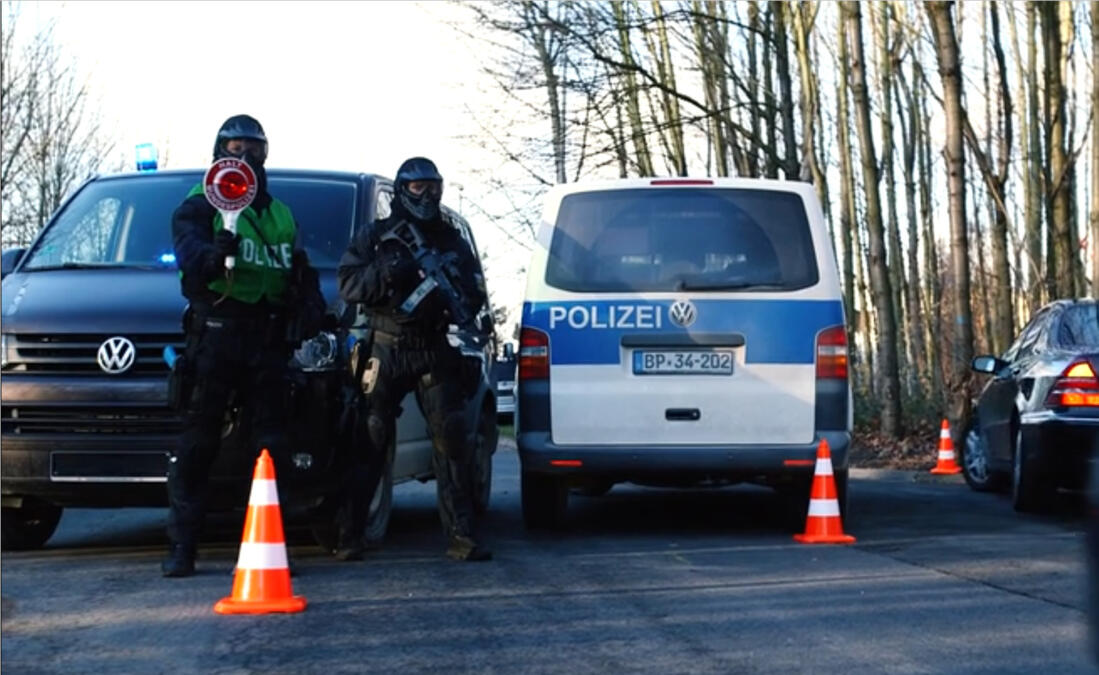 Das Bild zeigt eine Straßenkontrolle der Polizei, ein Beamter hält eine Polizeikelle, ein anderer ist vermummt und trägt eine automatische Waffe.