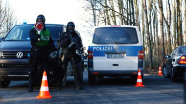 Das Bild zeigt eine Straßenkontrolle der Polizei, ein Beamter hält eine Polizeikelle, ein anderer ist vermummt und trägt eine automatische Waffe.