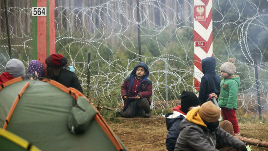 Stacheldrahtzaun hinter Grenz-Stelen von Polen, davor sitzen mehrere Kinder, im Vordergrund ein Zelt