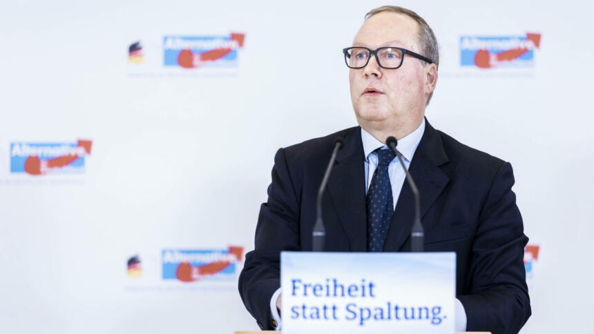 Ein Mann im schwarzen Anzug und mit schwarzer Brille am Rednerpult, davor der Slogan "Freiheit statt Spaltung", hinter ihm das Logo der AfD