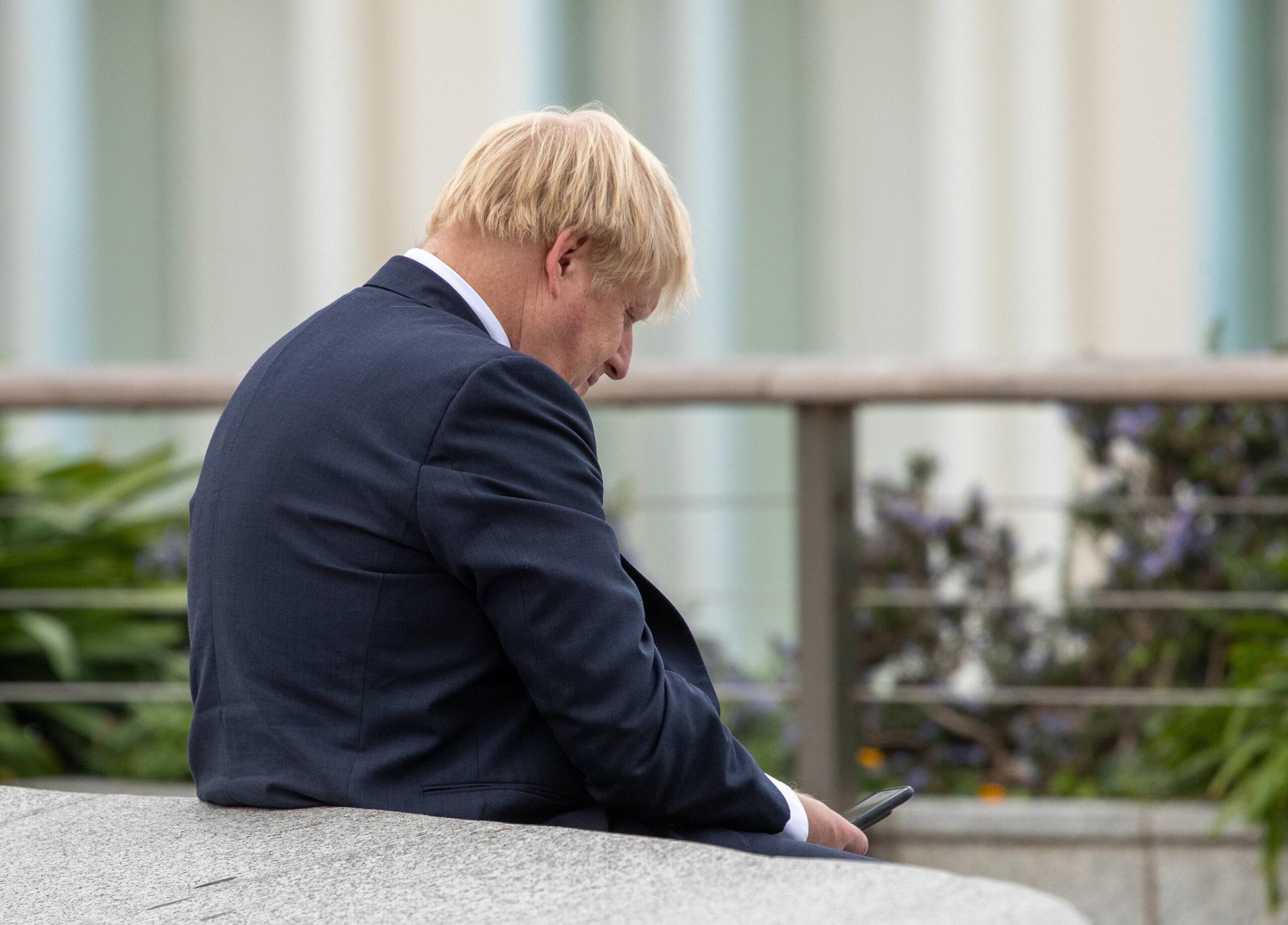 Boris Johnson lehnt sich an eine Mauer und schaut auf sein Smartphone
