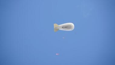 Auf dem Bild ist ein entferntes Luftschiff vor blauem Himmel zu sehen, an einer Leine hängen orangefarbene Gegenstände.