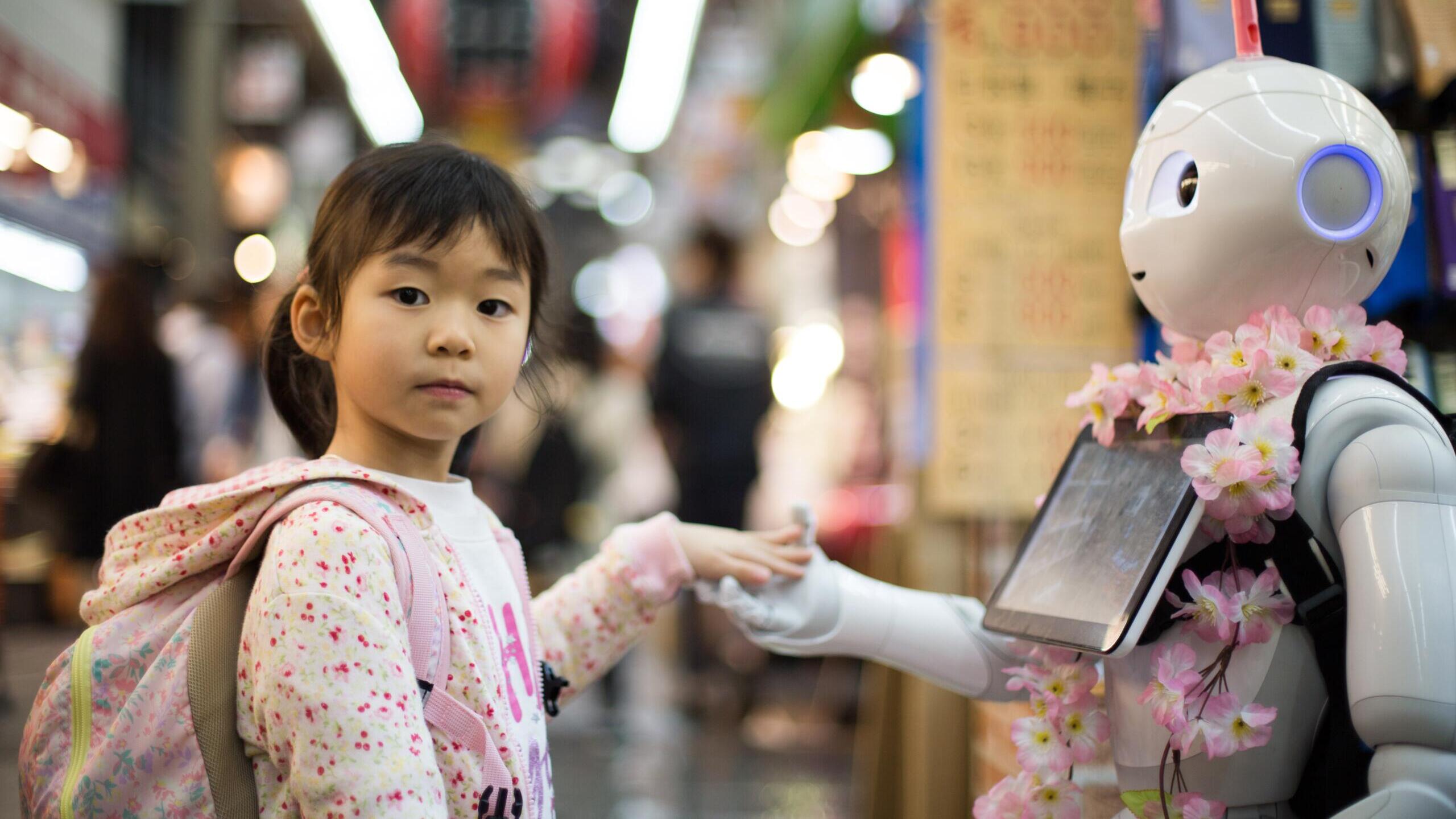 Kind gibt Roboter die Hand und schaut dabei in die Kamera