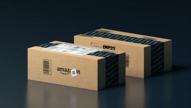 Zwei Amazon-Pakete liegen auf einem glänzenden Boden vor einem schwarzen Hintergrund.