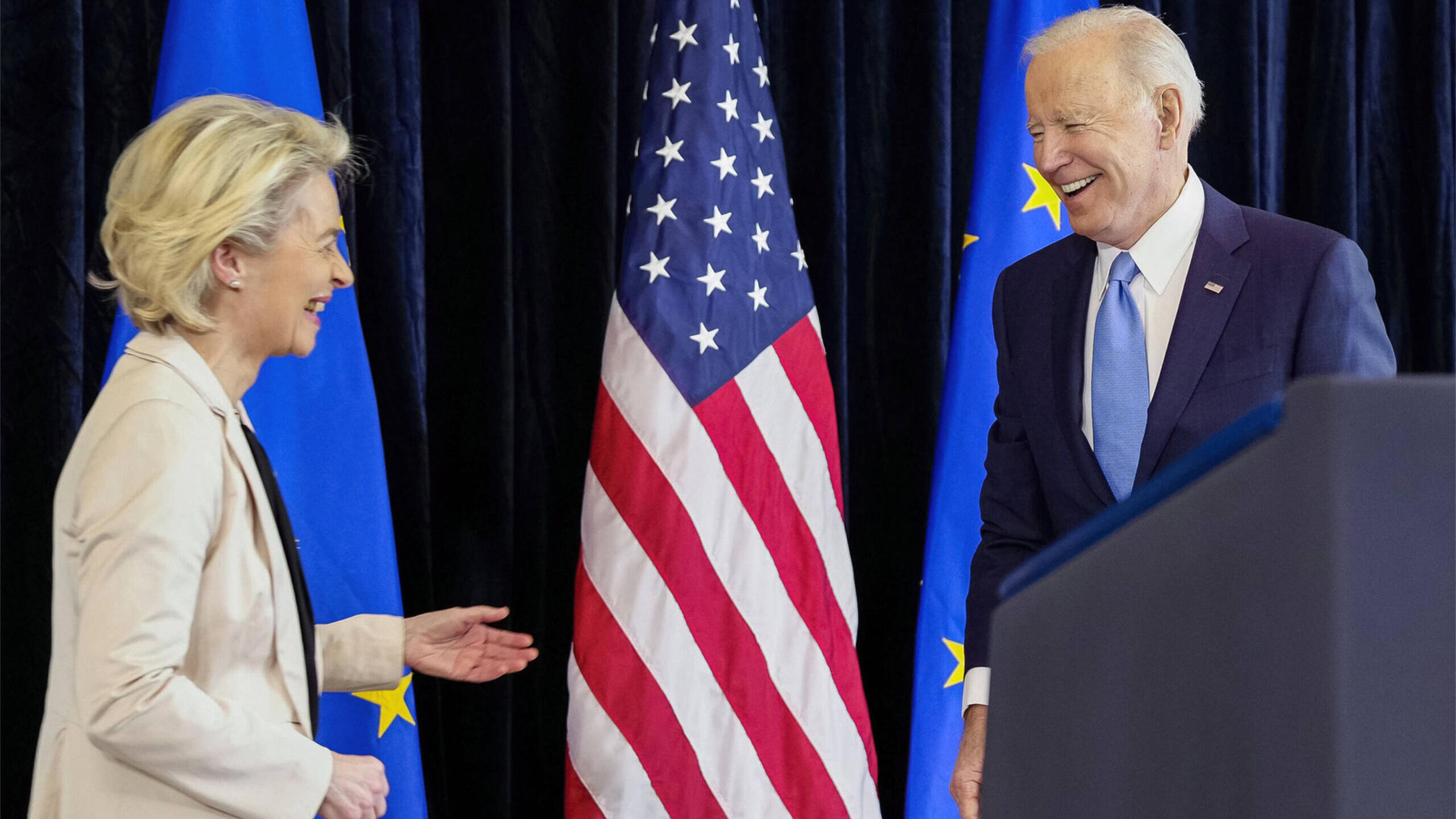 Eine Frau und ein Mann, lachend, vor Fahnen der USA und EU