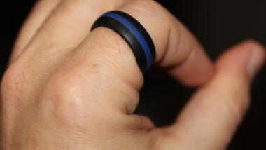 Das Bild zeigt eine linke Hand auf schwarzem Grund, am Zeigefinger befindet sich ein schwarz-blau-schwarz gestreifter Ring.