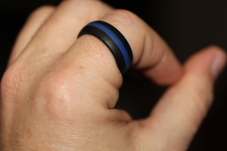 Das Bild zeigt eine linke Hand auf schwarzem Grund, am Zeigefinger befindet sich ein schwarz-blau-schwarz gestreifter Ring.