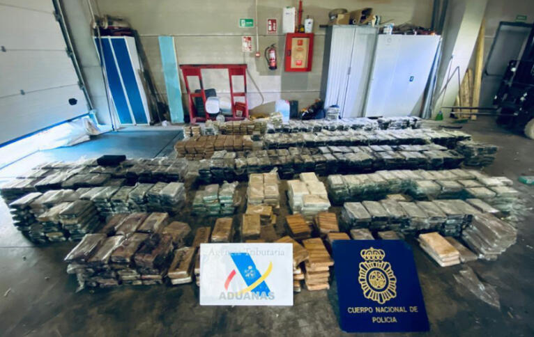 Das Bild zeigt sichergestellte Päckchen mit Rauschmitteln in einem Raum mit Schränken, davor sind zwei Logos von Zoll und Polizei aus Spanien montiert.