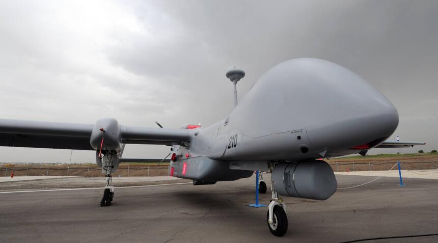 Die Drohne Heron TP steht auf einem Flugplatz. Im Hintergrund ist ein Funkturm zu sehen, es ist stark bewölkt.