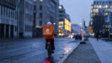 Eine Straße während der Dämmerung ist zu sehen. Man sieht einen Lieferando-Fahrer auf einem Fahrrad von hinten. Im Hintergrund sieht man verschwommen Autos und Häuser. Die Straße ist nass.