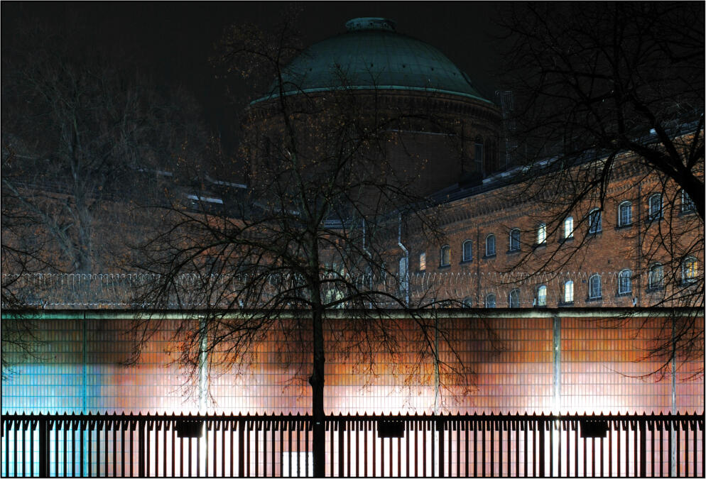 Die JVA Moabit bei Nacht. Im Vordergrund ist eine beleuchtete Mauer zu sehen. Auf der Mauer ist Staheldrahtzaun. Im Hintergrund ist das Gefängnis, in einigen Zellen brennt das Licht.