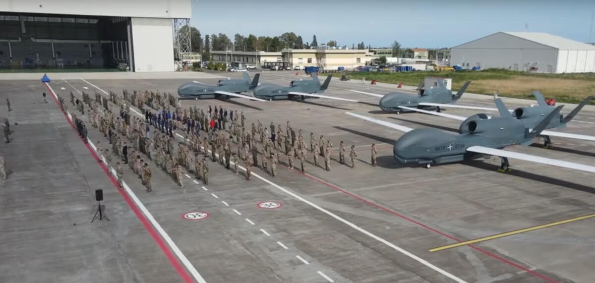 Ein Rollfeld mit 5 Global Hawk, davor stehen mehrere Dutzend Soldat:innen stramm, im Hintergrund ein Hangar.