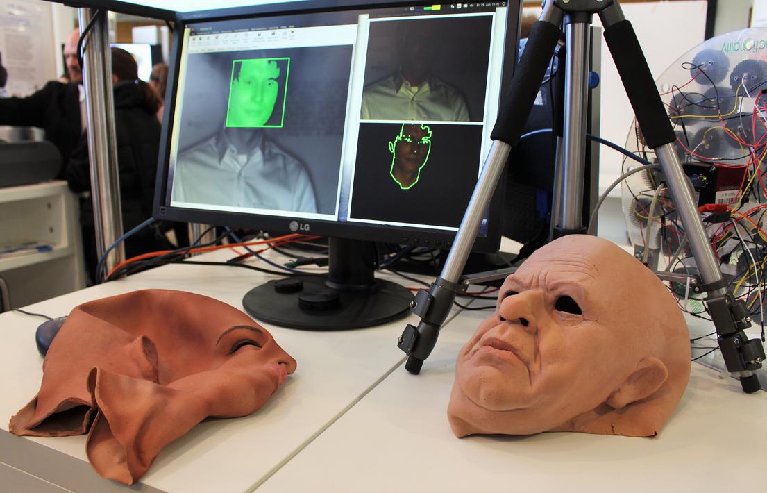 Vor einem Monitor, auf dem ein Gesichtserkennungsprogramm geöffnet ist, liegen zwei Gesichtsmasken.