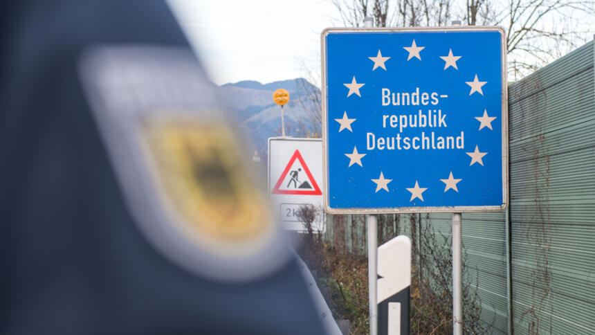 Das Bild zeigt ein Schild mit der Aufschrift "Bundesrepublik Deutschland", davor verscwommen ein Polizeiabzeichen an einem Ärmel.