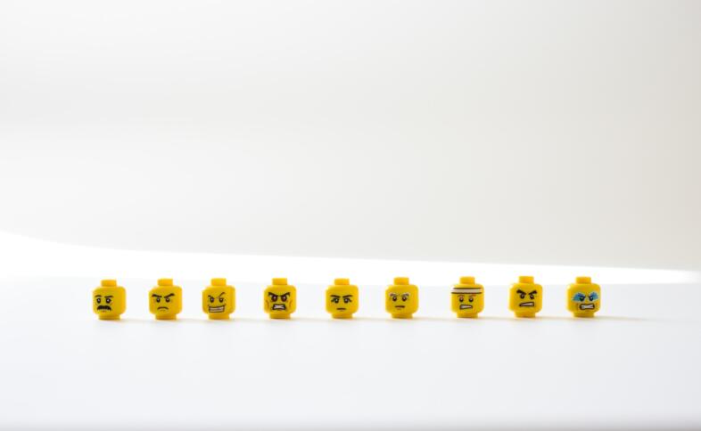 Auf einem weißen Tisch vor weißem Hintergrund sind 9 gelbe Lego-Köüfe zu sehen. Sie haben alle unterschiedliche Emotionen. Manche schauen wütend, andere Lachen, andere weinen oder sehen besorgt aus.