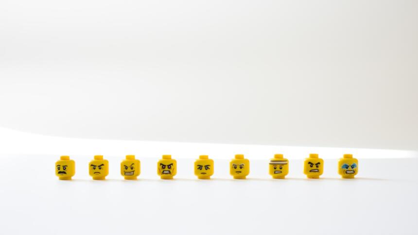 Auf einem weißen Tisch vor weißem Hintergrund sind 9 gelbe Lego-Köüfe zu sehen. Sie haben alle unterschiedliche Emotionen. Manche schauen wütend, andere Lachen, andere weinen oder sehen besorgt aus.