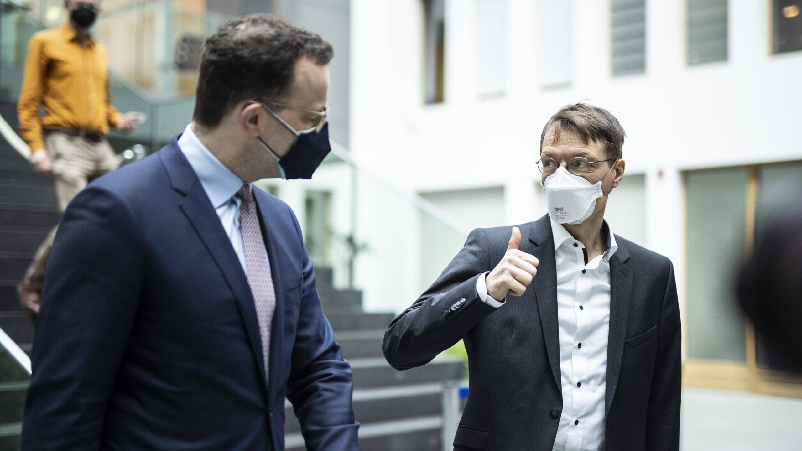 Im Vordergrund steht Jens Spahn, man sieht ihn von der Seite. Er guckt nach hinten rechts zu Karl Lauterbach, der einen Daumen nach oben zeigt und Spahn ansieht. Im Hintergrund ist ein Treppe. Beide tragen Maske