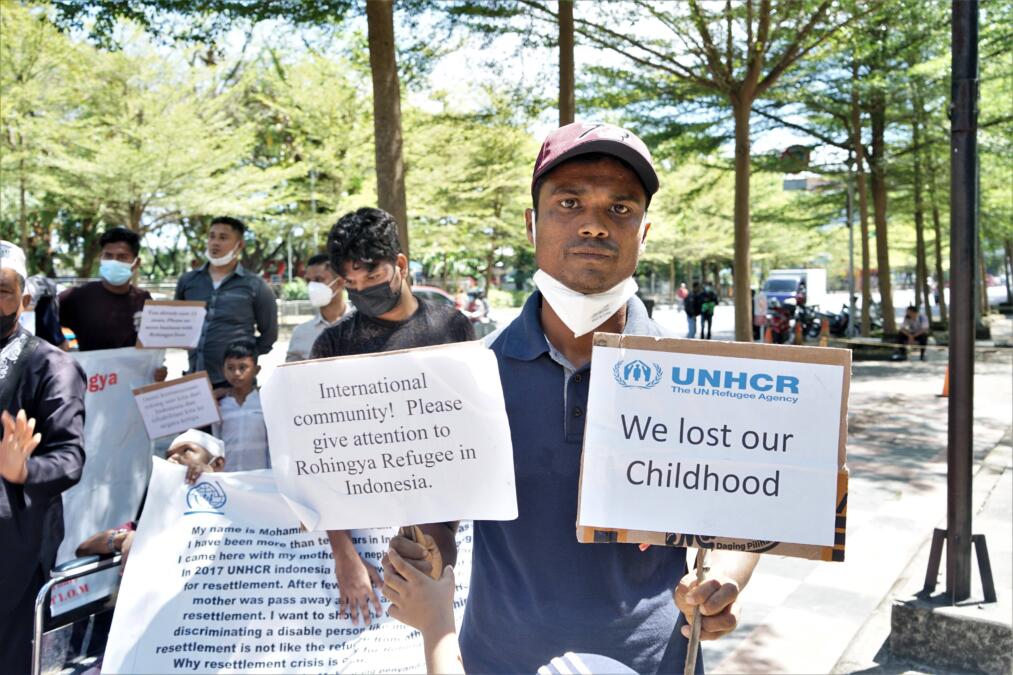 Eine Demonstration ist zu sehen. Im Vordergund des Bildes steht ein Mann. Er hält zwei Schilder hoch. Auf einem steht: "International Community! Please give attention to Rohingya Refugee in Indonesia." Auf dem anderen Schild steht: "We lost our Childhood". Im Hintergrund sind weitere Personen mit Schildern zu sehen.