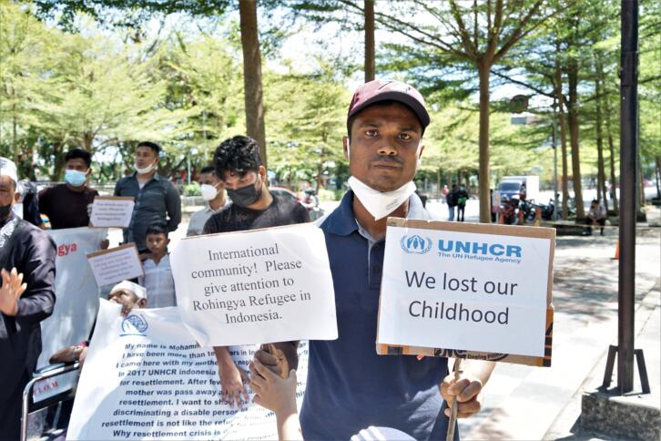 Eine Demonstration ist zu sehen. Im Vordergund des Bildes steht ein Mann. Er hält zwei Schilder hoch. Auf einem steht: "International Community! Please give attention to Rohingya Refugee in Indonesia." Auf dem anderen Schild steht: "We lost our Childhood". Im Hintergrund sind weitere Personen mit Schildern zu sehen.