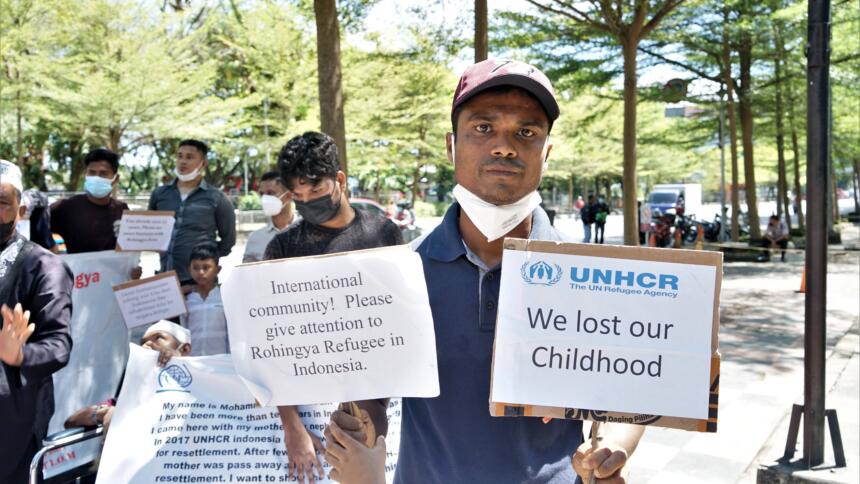 Eine Demonstration ist zu sehen. Im Vordergund des Bildes steht ein Mann. Er hält zwei Schilder hoch. Auf einem steht: "International Community! Please give attention to Rohingya Refugee in Indonesia." Auf dem anderen Schild steht: "We lost our Childhood". Im Hintergrund sind weitere Personen mit Schildern zu sehen.