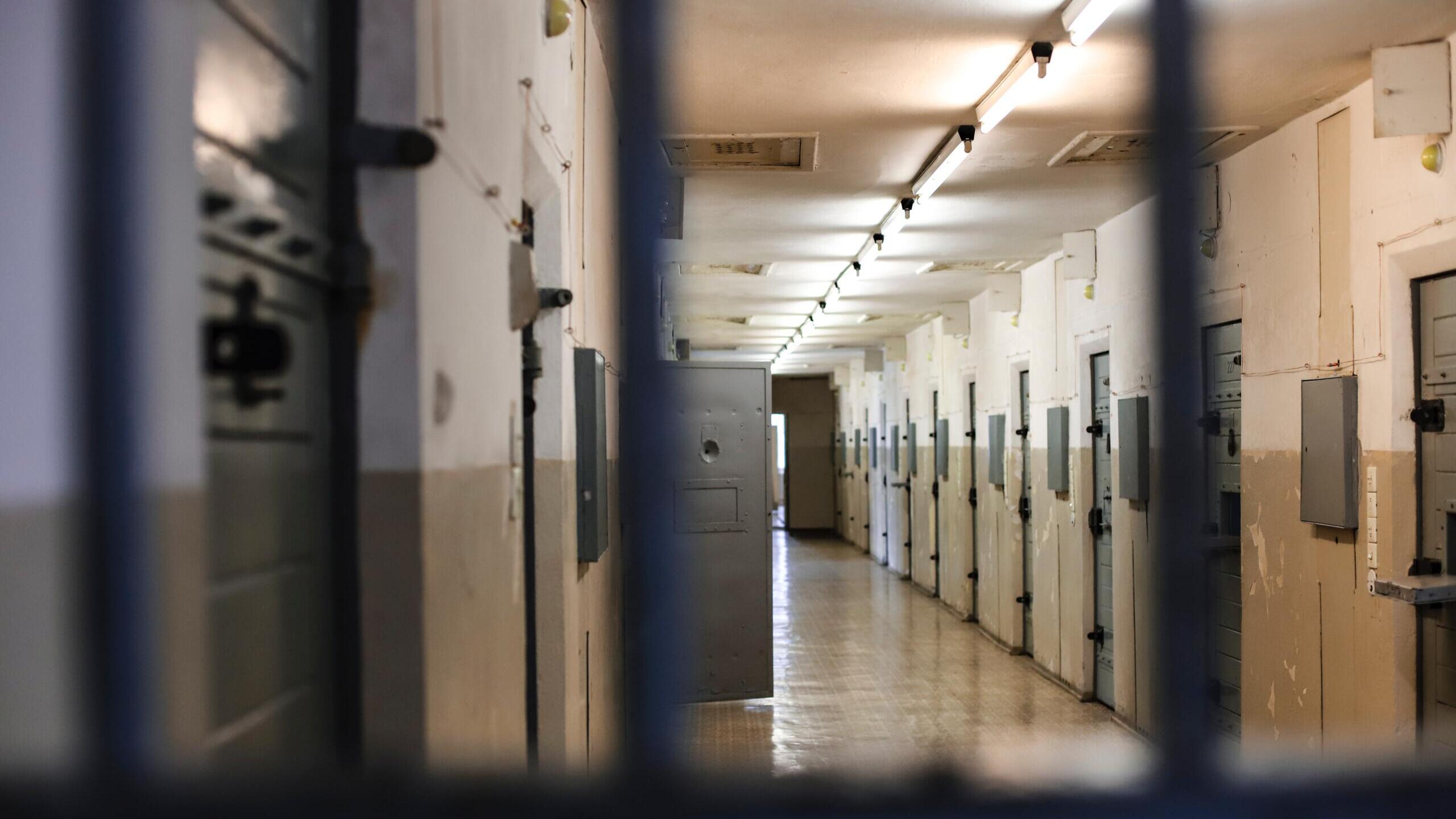 Im Vordergrund sind verschwommen Gitterstäbe zu sehen. Im Hintergrund ist einhellerleuchteter Gefängnistrakt mit Zellentüren zu sehen