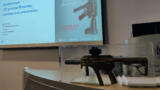 Im Hintergrund eine Präsentation zu gedruckten feuerwaffen, im Vordergrund eine Maschinenpistole.