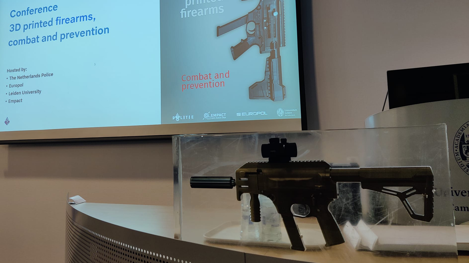 Im Hintergrund eine Präsentation zu gedruckten feuerwaffen, im Vordergrund eine Maschinenpistole.