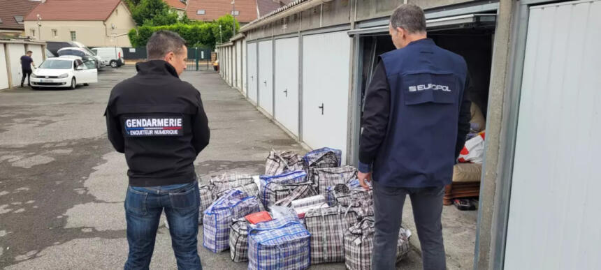 Zwei Polizisten der frz. Gendarmerie und von Europol stehen mit dem Rücken zur Kamera vor einer offenen Garage. Vor ihnenstehen auf dem Boden zahlreiche große Taschen.