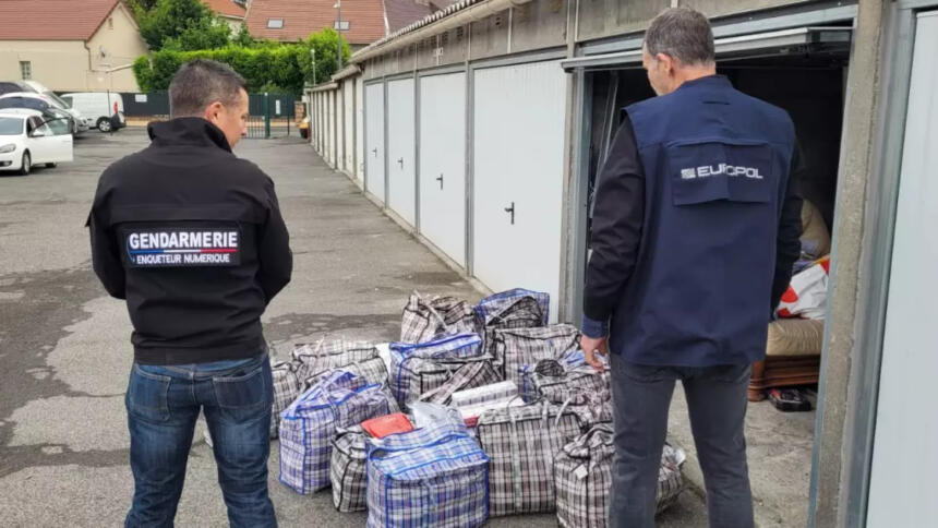 Zwei Polizisten der frz. Gendarmerie und von Europol stehen mit dem Rücken zur Kamera vor einer offenen Garage. Vor ihnenstehen auf dem Boden zahlreiche große Taschen.