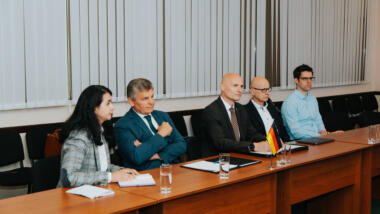 Vier Männer und möglicherweise eine Übersetzerin sitzen in einer Reihe und schauen konzentriert, vor ihnen eine Deutschlandfahne.