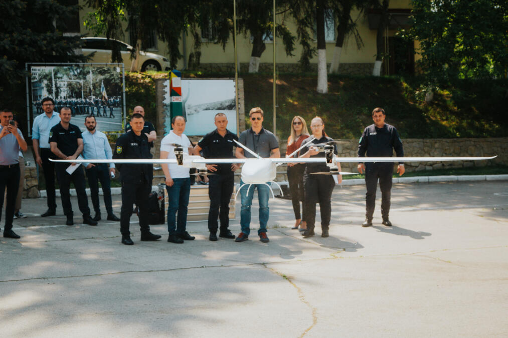Eine Gruppe aus mehreren Männern und zwei Frauen steht im Hintergrund, während einer von diesen eine Starrflügler-Drohne steuert die im Vordergrund senkrecht abhebt.