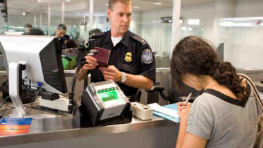 Ein Grenzbeamter blickt argwöhnsich auf eine reisende, deren Pass er in den Händen hält, in der Bildmitte ein Fingerabdruckscanner.