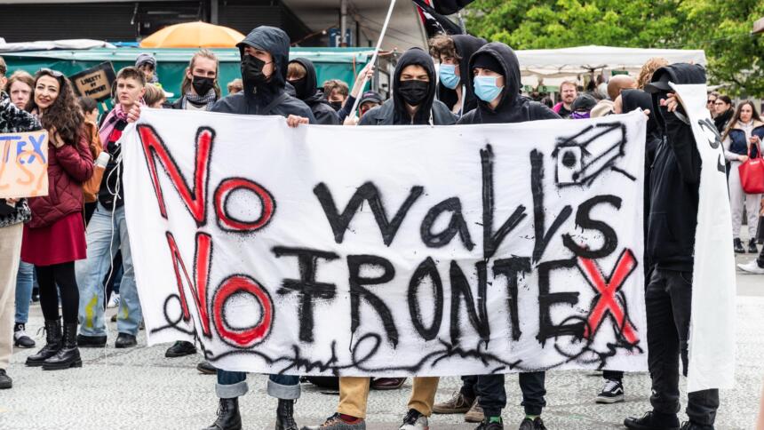 drei Demonstranten mit Masken stehen hinter einem weißen Banner, auf dem steht: No walls, no Frontex