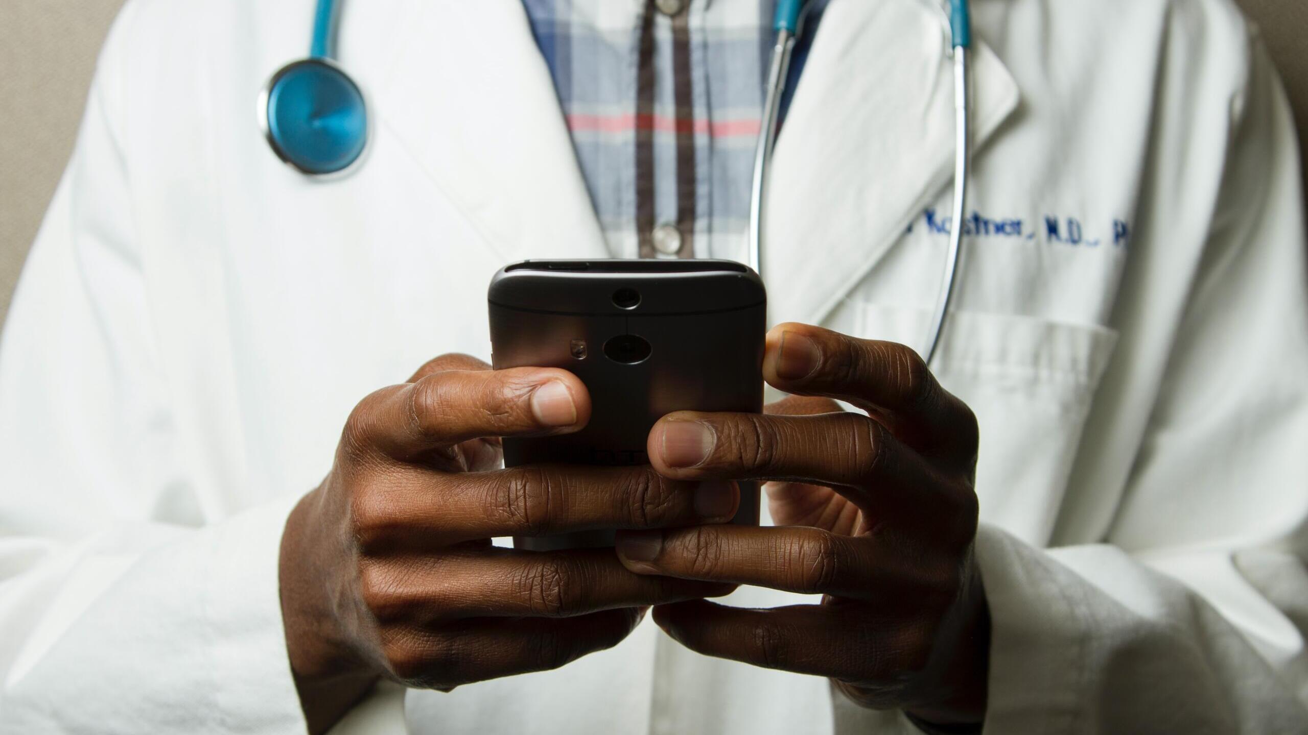 ein arzt mit weißem kittel und stethoskop um den hals hält ein handy in der hand