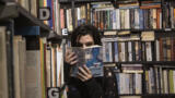 Eine Frau steht zwischen Bücherregalen und schaut über den Buchrücken in die Kamera.