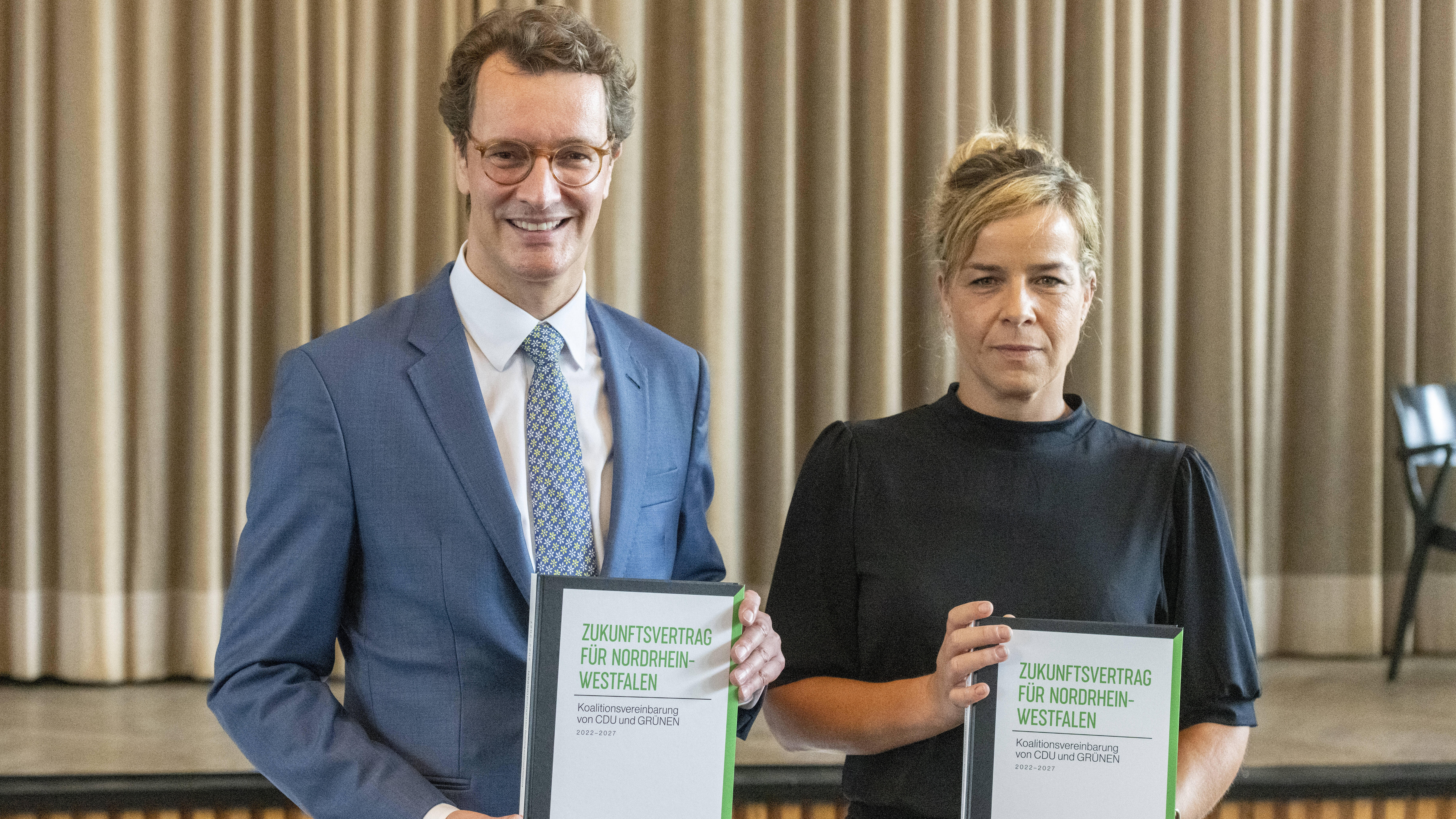 Hendrik Wüst und Mona Neubaur stehen nebeneinander und halten den Koalitionsvertrag in die Kamera