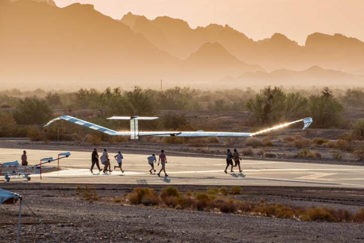 Das Luftfahrzeug im Flug über einer Startbahn, darunter mehrere Personen, im Hintergrund Berge und Dunst.