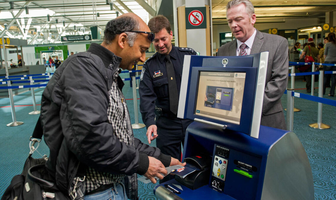 Ein Selbstbedienungskiosk in der rechten Bildhälfte, in die ein Reisender einen Ausweis einführt, beobachtet von zwei freudigen Beamten.