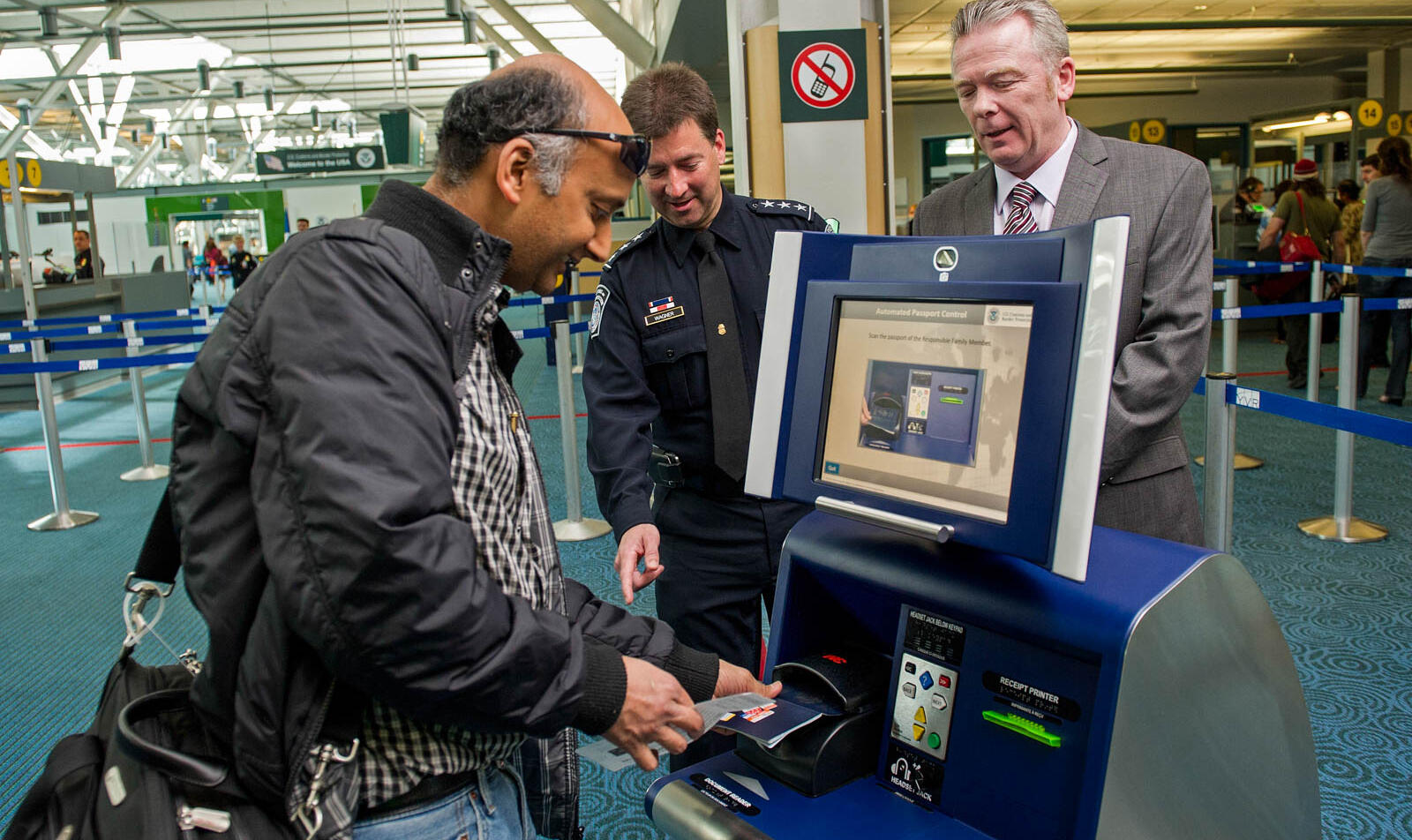 Ein Selbstbedienungskiosk in der rechten Bildhälfte, in die ein Reisender einen Ausweis einführt, beobachtet von zwei freudigen Beamten.