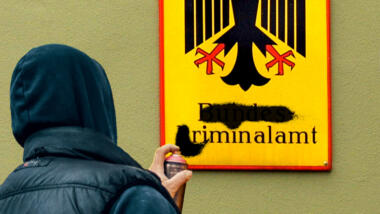 Ein Graffitisprayer sprüht den Schriftzug Bundeskriminalamt auf dem Behördenschild an (Fotomontage)