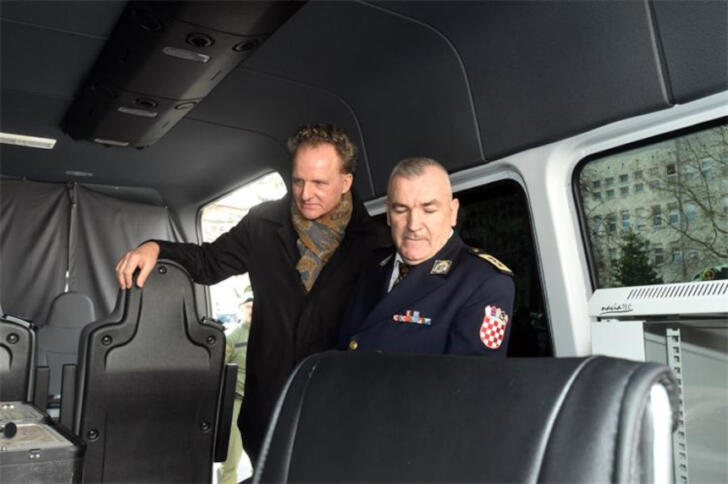 Zwei Männer, davon einer in Uniform, im Innenraum eines schwarz ausgekleideten Fahrzeugs.