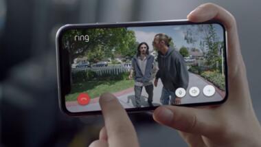 Großaufnahme eines Smartphones, auf dem Bilder aus einer Überwachungskamera mit zwei Männern in einem Vorgarten zu sehen
