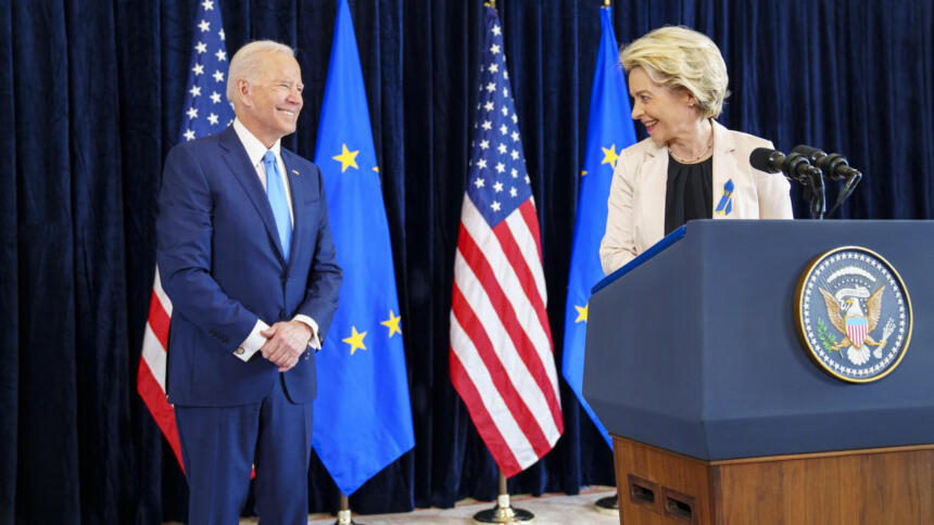 Im Hintergrund EU- und USA-Flaggen, im Vordergrund: Links ein Mann mit weißen Haaren im Anzug, rechts eine blonde Frau am Rednerpult, die ihm den Kopf zudreht