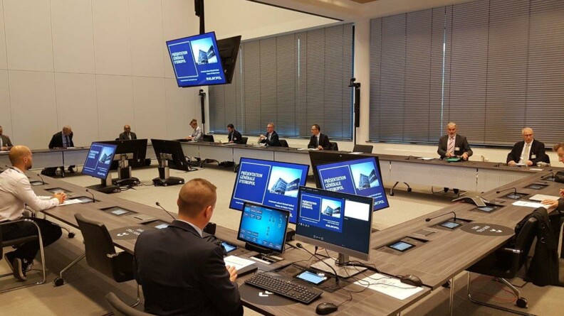 MehrerePersonen sitzen in einem Tischkreis vor Monitoren, darauf zu erkennen ist das Gebäude von Europol.